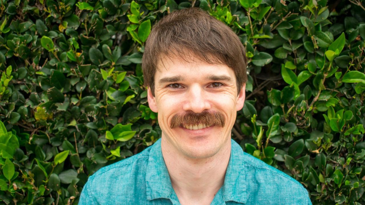 A man, wearing a blue shirt, with a rich moustache and smile on hisface is standing in front of a thick, bright green hedge.