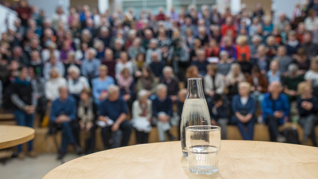 Bild på vattenkaraff med glas på ett bord framför en publik. Bilden ska illustrera att hålla föredrag.