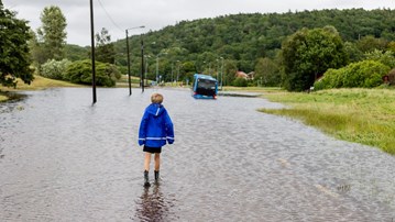 Ett barn står vänd mot en översvämmad bilväg. Längre fram syns en buss.