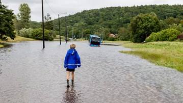 Ett barn står vänd mot en översvämmad bilväg. Längre fram syns en buss.