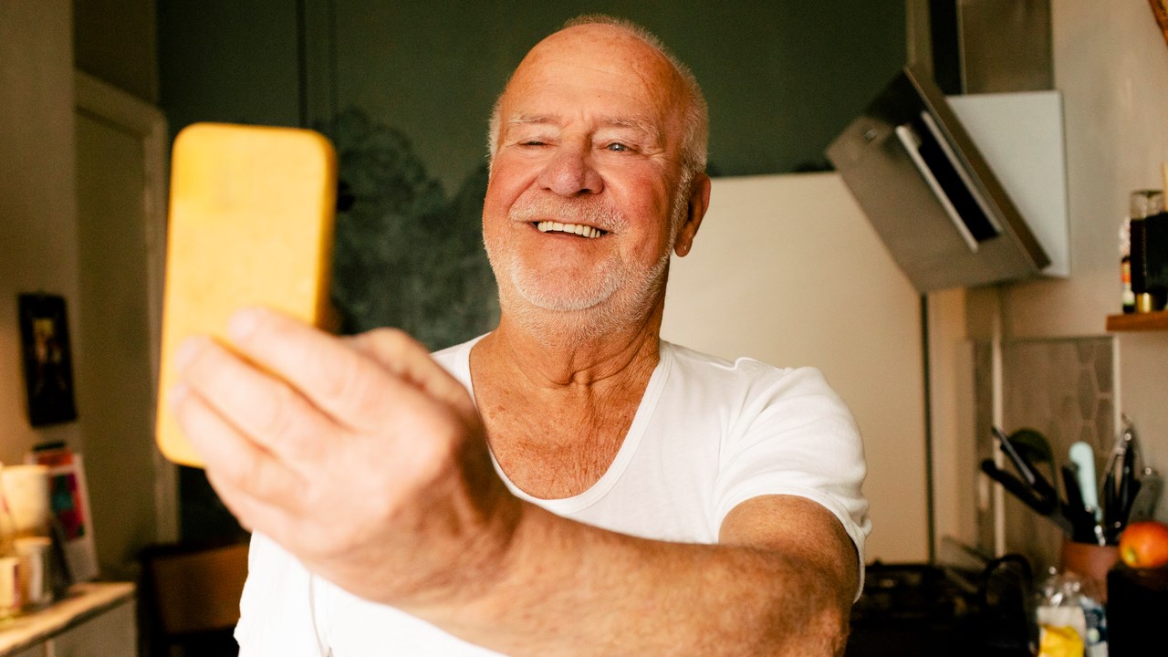 En glad äldre flintskallig man med lätt skäggstubb och vit tröja står i kök och tittar i sin mobiltelefon