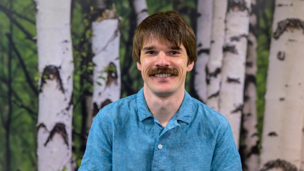 A man with a mustache and a blue shirt poses in front of a birch tree background