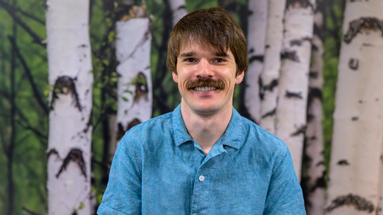 A man with a mustache and a blue shirt poses in front of a birch tree background