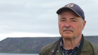 Bild på Lennart Bondesson som utomhus med en sjö och berg i bakgrunden. Han bär en keps och en grön jacka och ser direkt in i kameran.