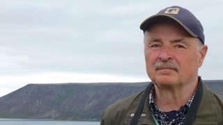 Bild på Lennart Bondesson som utomhus med en sjö och berg i bakgrunden. Han bär en keps och en grön jacka och ser direkt in i kameran.