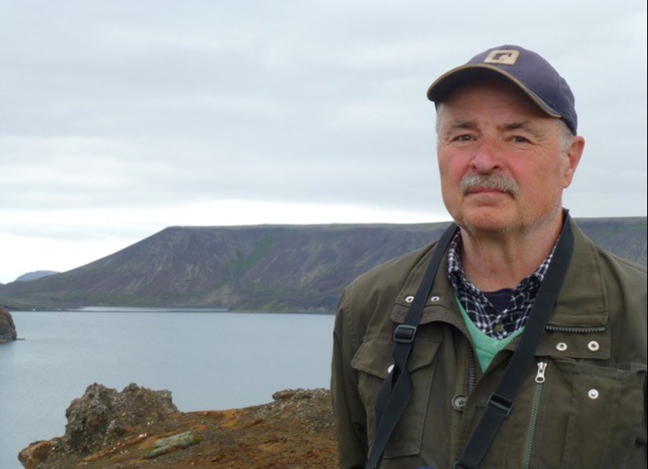 Bild på Lennart Bondesson som utomhus med en sjö och berg i bakgrunden. Han bär en keps och en grön jacka och ser direkt in i kameran.