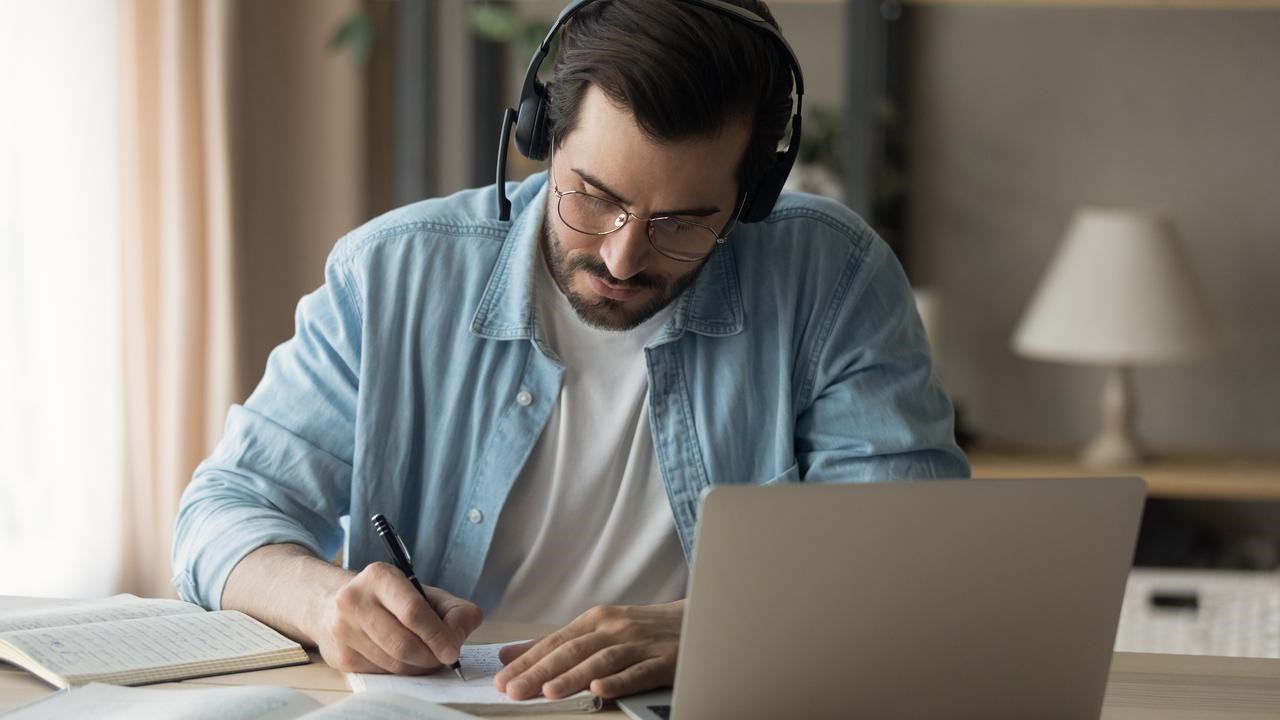 En man sitter vid ett bord med laptop och headset, böcker och antecknar i ett block.