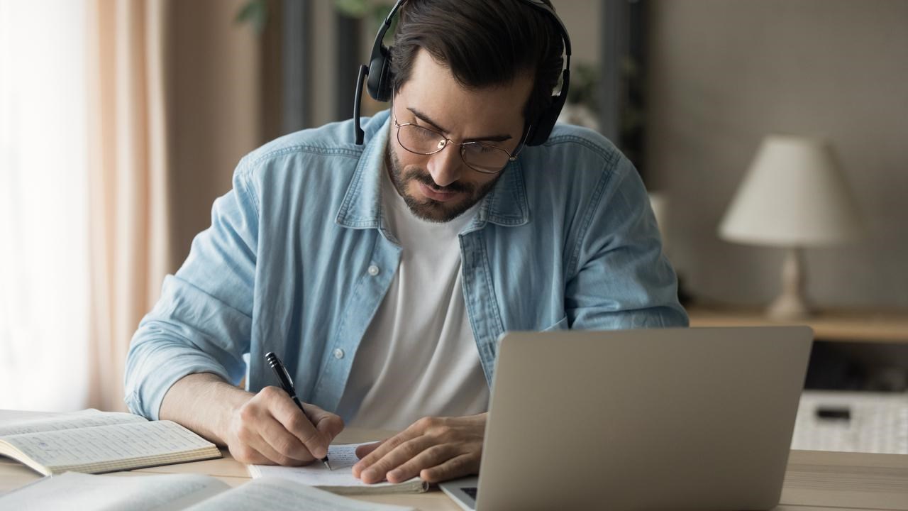En man sitter vid ett bord med laptop och headset, böcker och antecknar i ett block.