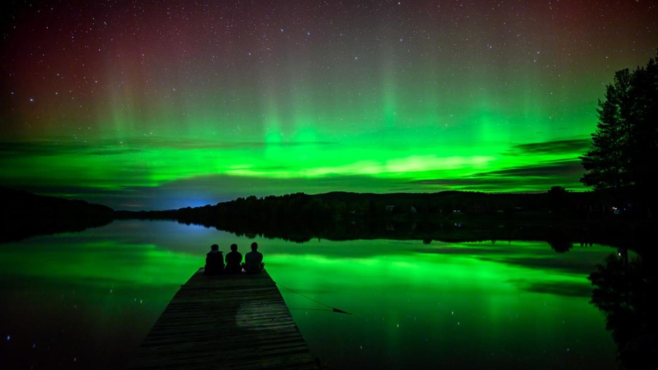 A group of people sitting on a dock watching the aurora reflecting in a river.