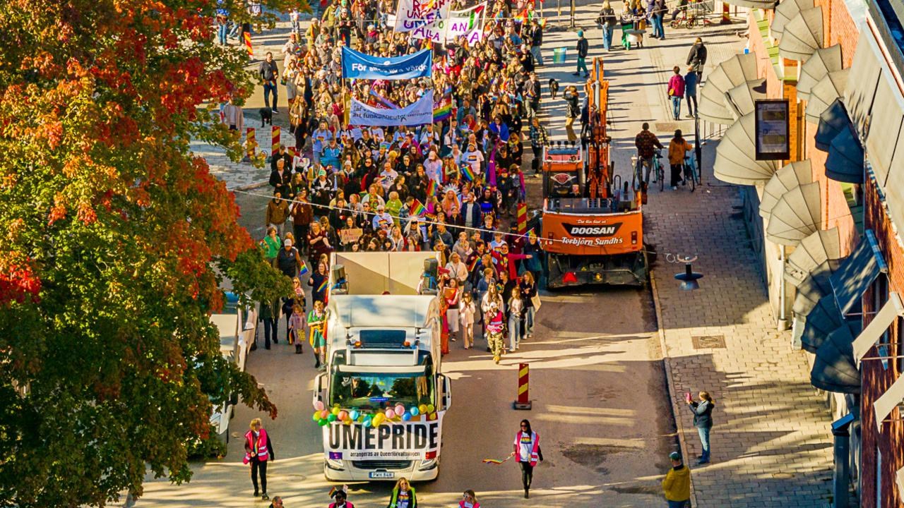 Drönarbild av en parad av människor som går i Pride-tåget i Umeå 2024, med regnbågsflaggor och banderoller.