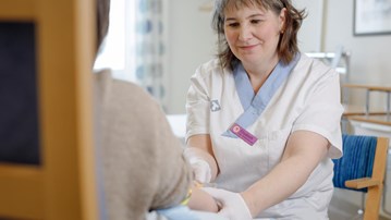 En person sitter med ryggen mot kameran och håller fram armen. Mitt emot sitter studiekoordinatorn Elisabeth Müller Granberg i färd med att ta ett blodprov.