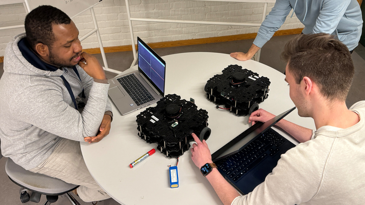 ree students sit down around a round table. They have computers and two robots. They are discussing, behind them there is a board with mathematical calculations.