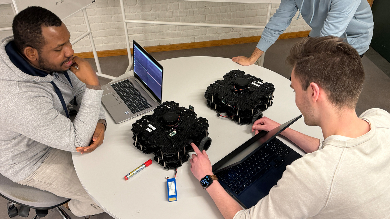 Tre studenter sitter ned kring ett runt bord. De har datorer och två robotar på hjul. De diskuterar, bakom dem finns det en tavla med matematiska beräkningar.