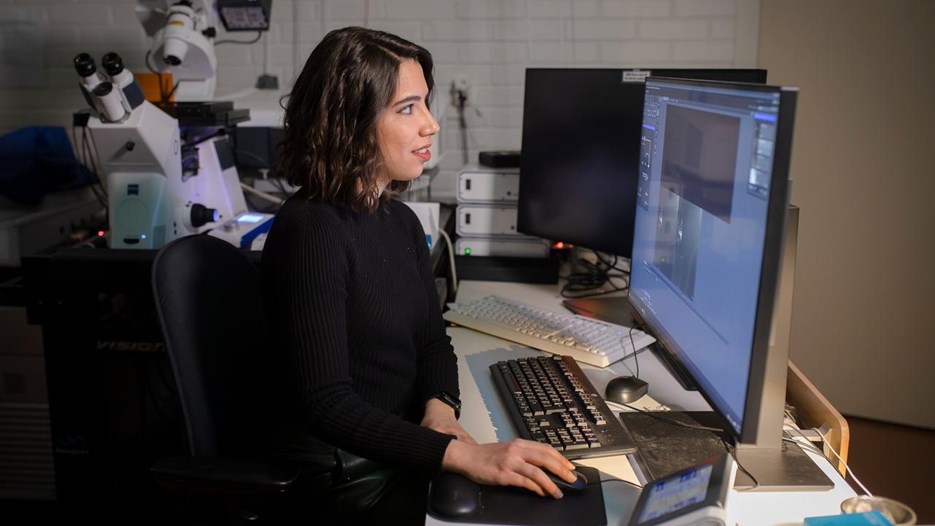A woman with medium-length brown hair sits in front of a computer screen connected to an advanced microscope.