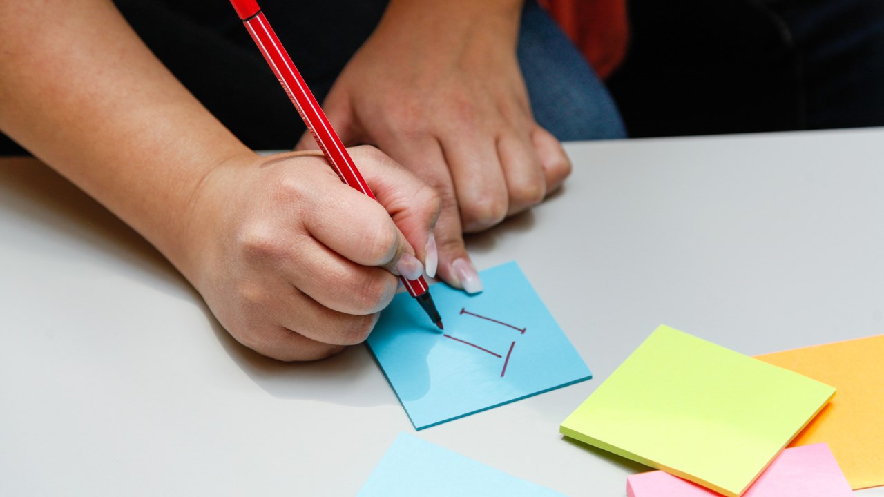 Hand som skriver IT på en post-it-lapp.