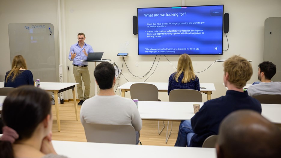 A group of researchers are seated at white tables with their backs to the camera. They face Anders Garpebring who stands by a round table with his laptop. The screen next to him shows a slide that says "What am I looking for?" followed by more text.