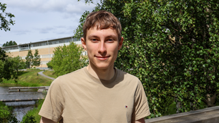 Person i ljusbeige t-shirt står vid ett trästaket utomhus. Ibakgrunden syns grönska, träd, en smal vattenkanal med bryggor samt en större byggnad. Soligt väder med inslag av moln på himlen.