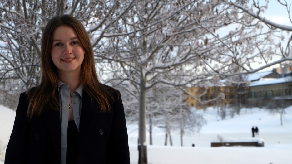 Bild på Elsa Karlsson Gustafsson, utomhus med en suddig bakgrund med snötäckta träd bakom samt universitets dammen med is på