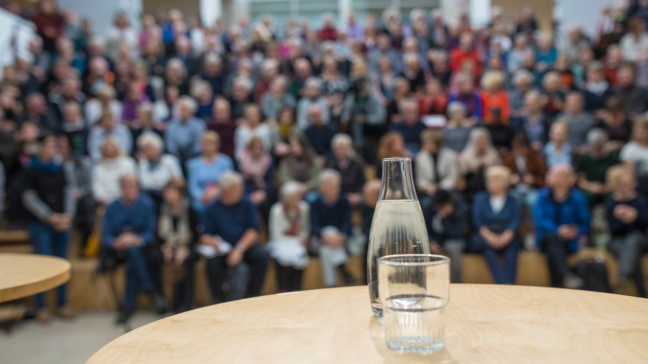 Bild på vattenkaraff med glas på ett bord framför en publik. Bilden ska illustrera att hålla föredrag.