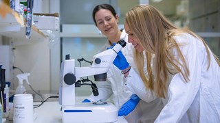 Två kvinnor i labbrockar tittar genom ett mikroskop