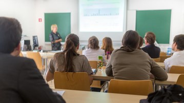 Föreläsningssal på universitet med studenter i förgrunden och en lärare vid tavlan.