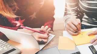 Två personer som sitter med datorer och block och skriver. Grupparbete.