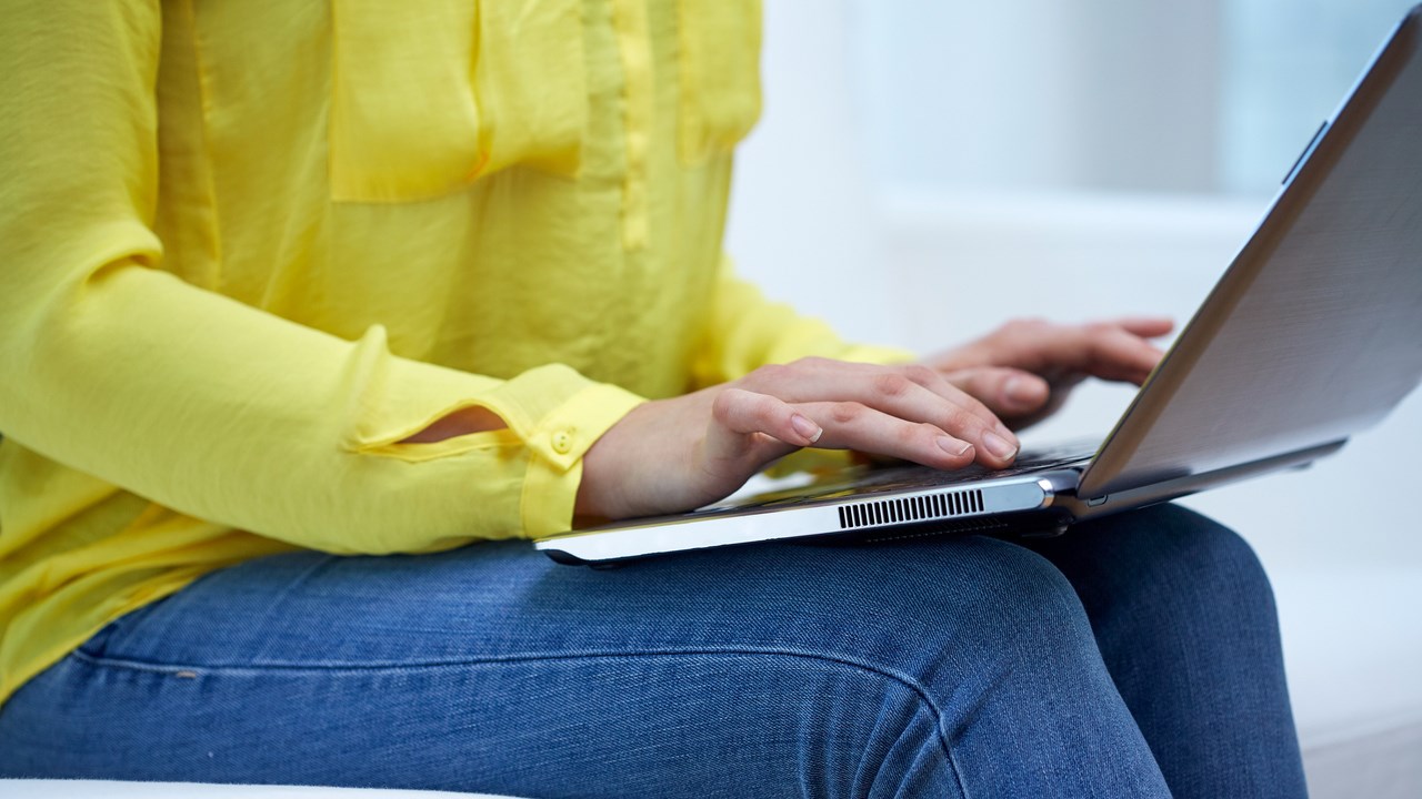 Detaljbild på kvinna i soffa som sitter med uppslagen laptop i en soffa och skriver.
