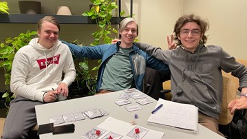Tre personer som sitter vid ett bord och ser glada ut, framför dem ligger tärningar och en prototyp av ett kortspel.
