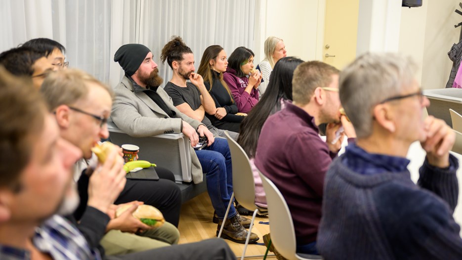 An audience of seated researchers listen with intent faces to a research pitch while some of them eat sandwiches