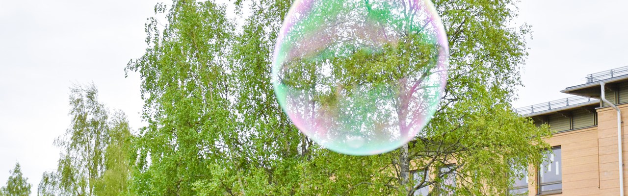 Giant soap bubble is flying in front of birch trees and a yellow brick building