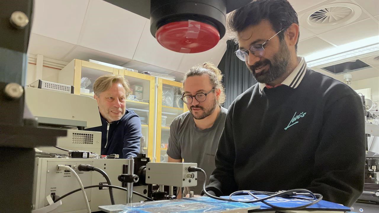 Tre män i ett laboratorium som tittar på något som ligger på ett bord.