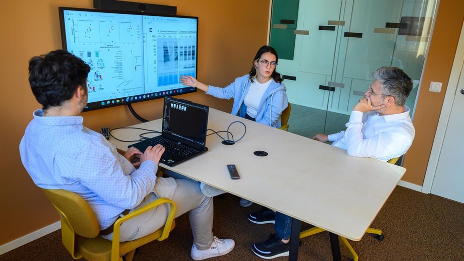 A group of people sitting around a table with laptops have a meeting and discuss research results on a screen.