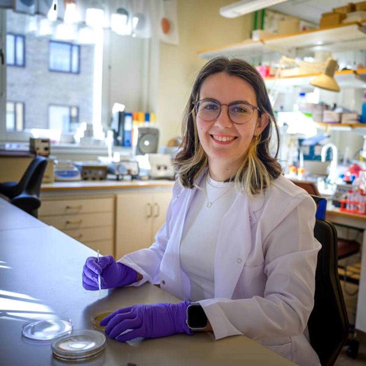 A woman in a lab coat and purple gloves holds a petri dish in one hand and a glass implement in her other hand.