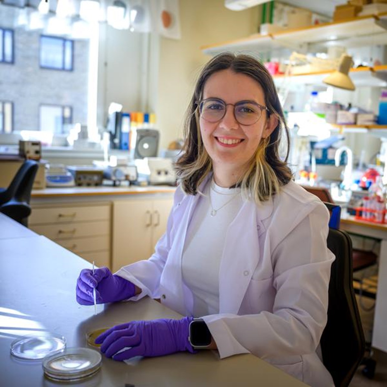 A woman in a lab coat and purple gloves holds a petri dish in one hand and a glass implement in her other hand.
