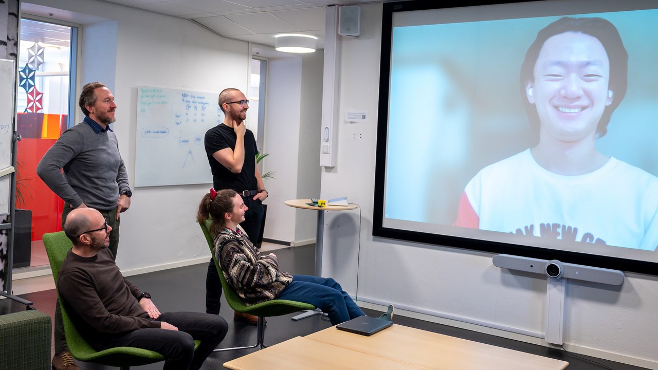 Four people, two seated and two standing, have a meeting with a smiling man on a screen