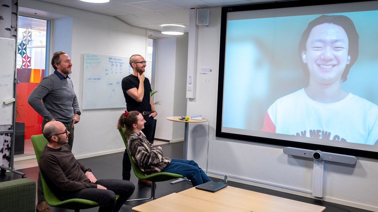 Four people, two seated and two standing, have a meeting with a smiling man on a screen