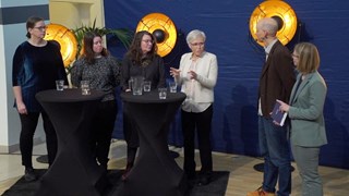 Fyra kvinnor står bredvid varandra vid ståbord och två moderatorer står vid sidan av. 