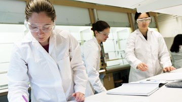 Bild på tre studenter i labbrockar som gör anteckningar i labbrapporter.