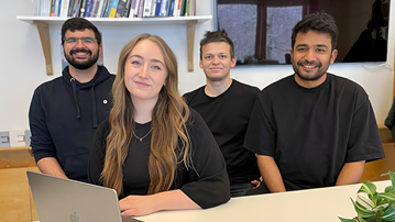 Bilden visar fyra personer, tre män och en kvinna som sitter vid ettbord med en dator framför sig.