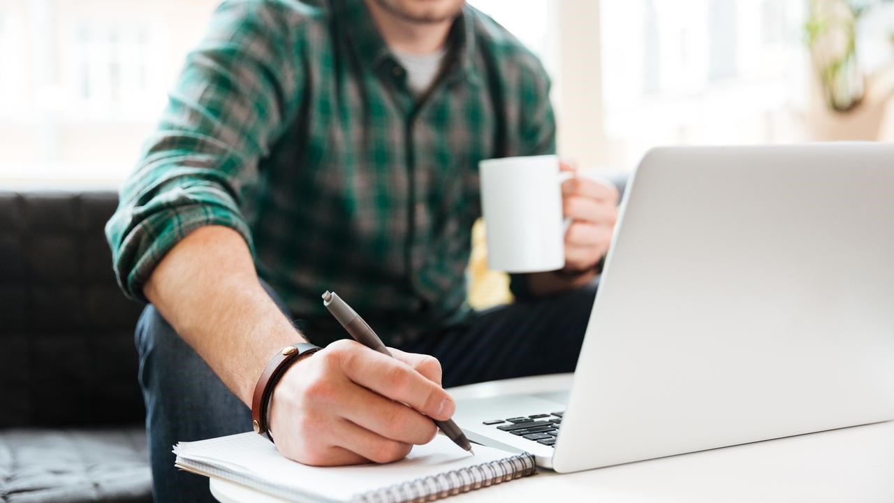 Person sitter i en soffa och arbetar. Har dator, kaffekopp och block där han skriver.