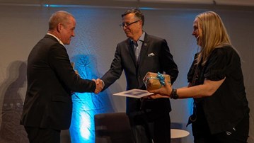 Dag Avango och Dag Rune Olsen står skakar hand. Bredvid dem står Johanne Raade med en present i handed. Alla är fint klädda i mörka kläder.