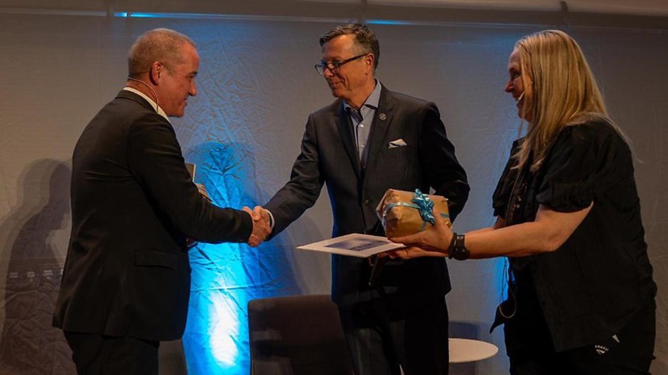Dag Avango och Dag Rune Olsen står och skakar hand. Bredvid dem står Johanne Raade med en present i handen. De är alla fint klädda i mörka kläder.