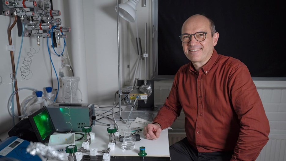 En man i röd skjorta och glasögon sitter i ett mörkt rum bredvid ett bord med teknisk utrustning. Han ler mot kameran.