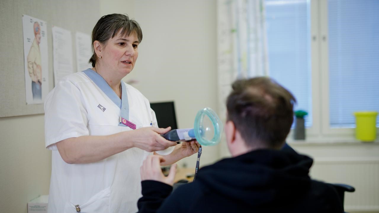 På ett undersökningsrum ses forskningssjuksköterskan Elisabeth Müller Granberg hålla fram en spirometer framför ALS-patienten som sitter i en stol med ryggen mot kameran och sträcker sig efter den.