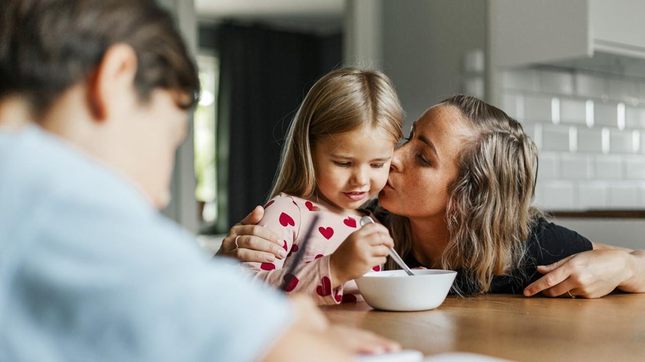 Mamma kysser barn på kinden vid frukoststund. En man syns i förgrunden.