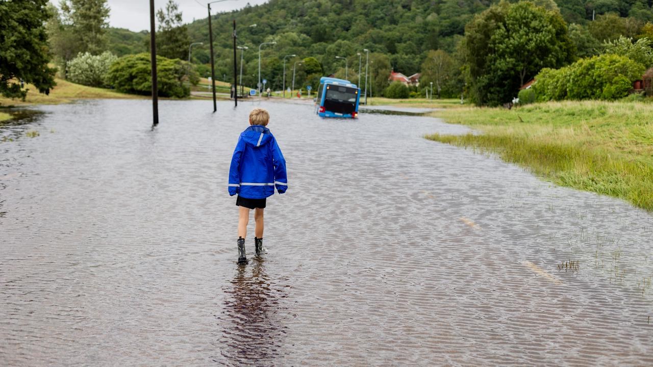 Ett barn står vänd mot en översvämmad bilväg. Längre fram syns en buss.