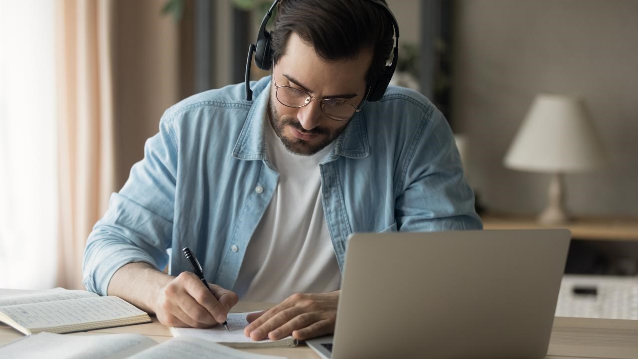 En man sitter vid ett bord med laptop och headset, böcker och antecknar i ett block.