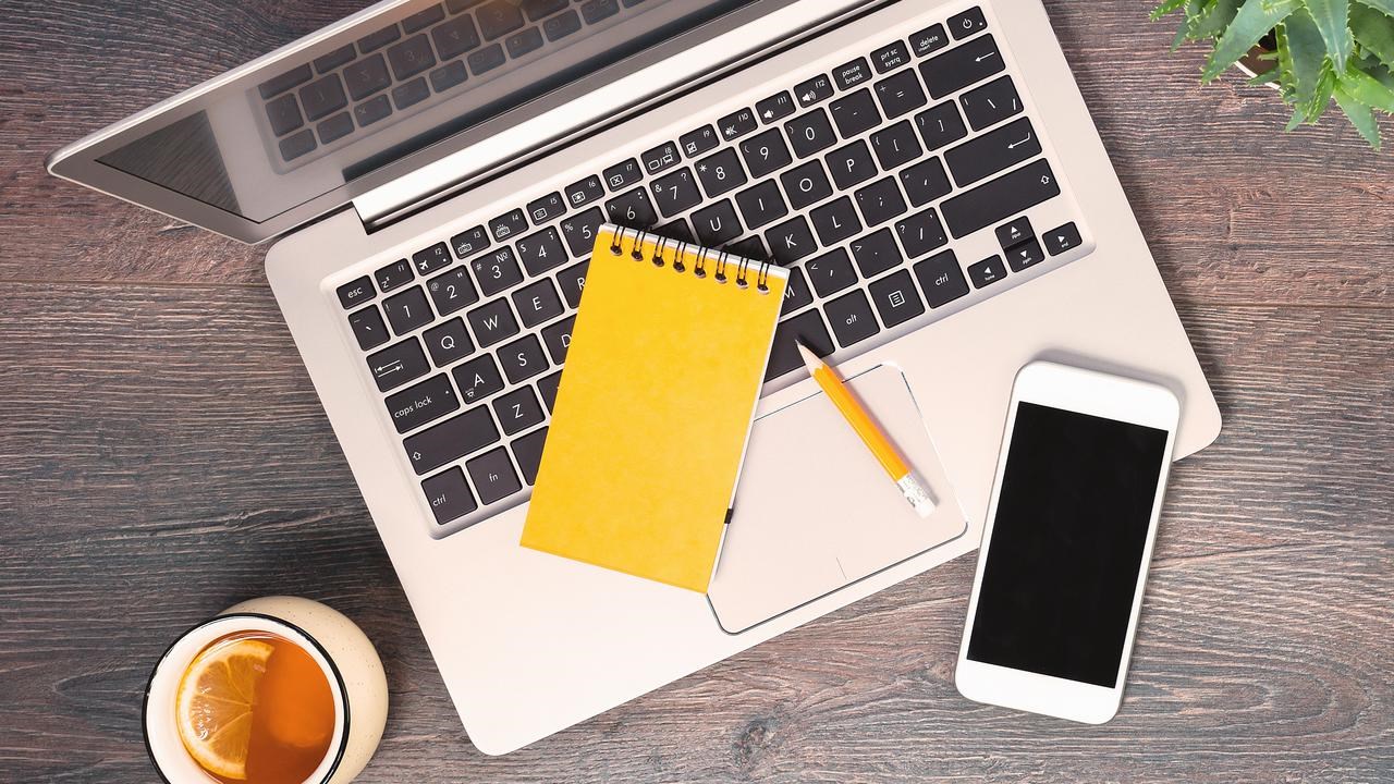 Dator, anteckningsblock, mobil och penna fotat uppifrån på ett bord.