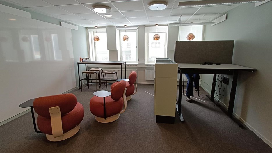 Open office with two desks to the right, and three red chairs to the left. The room is light with red hanging lights in the windows.