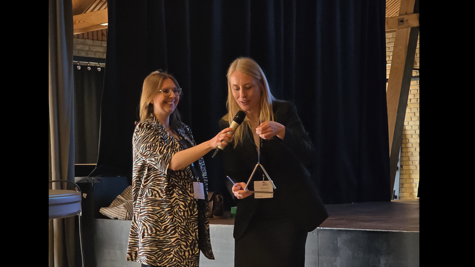 Värdarna för DHNB 2026, här representerade av Katrine L. Baunvig och Anne Agersnap, Århus universitet. De klingar ut konferensen och överlämnar därefter den ceremoniella triangeln i Umeå universitets vård.